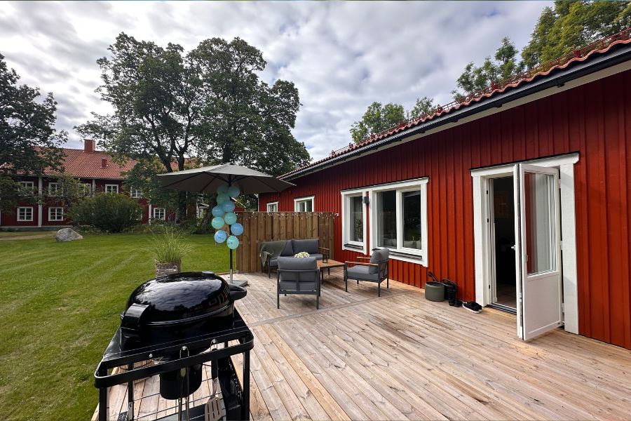 Röd svensk stuga med träterrass i soligt väder, utrustad med grill och möbler, omgiven av grönskande trädgård och träd