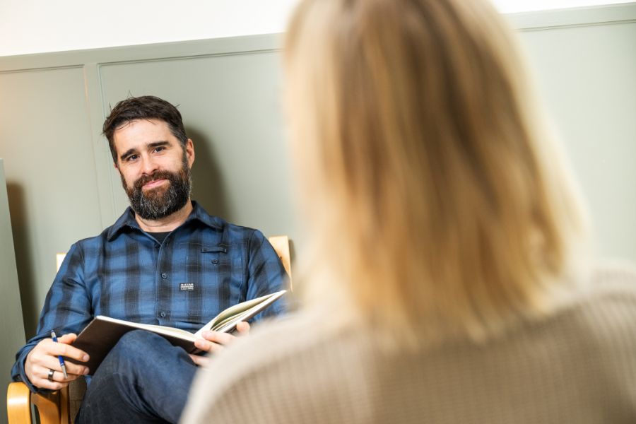 Man i blå skjorta som håller en anteckningsbok och sitter mittemot en kvinna i beige tröja under ett samtal i ett ljust rum