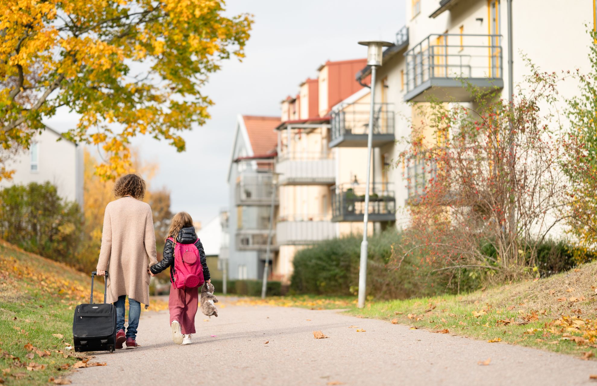 Kvinna med rullväska går hand i hand med en liten flicka med ryggsäck och gosedjur i handen