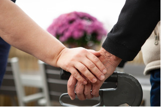 Två händer håller i en rollator utomhus, med suddiga rosa blommor i bakgrunden, symboliserar stöd och omsorg