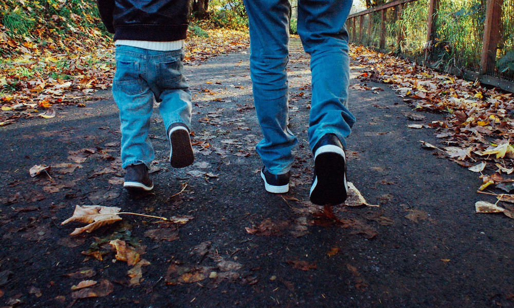 Ett barn går tillsammans med en vuxen på asfalterad väg täckt med höstlöv, båda iklädda jeans och sneakers
