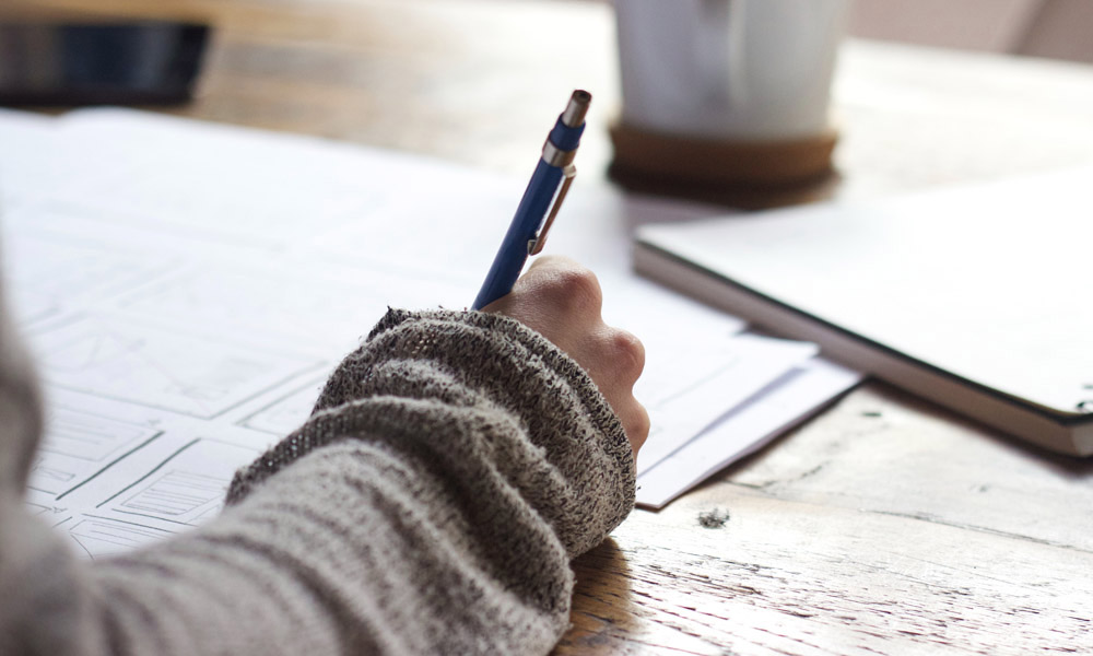 Elev skriver anteckningar med en penna vid ett träbord, papper och en böcker synliga i bakgrunden