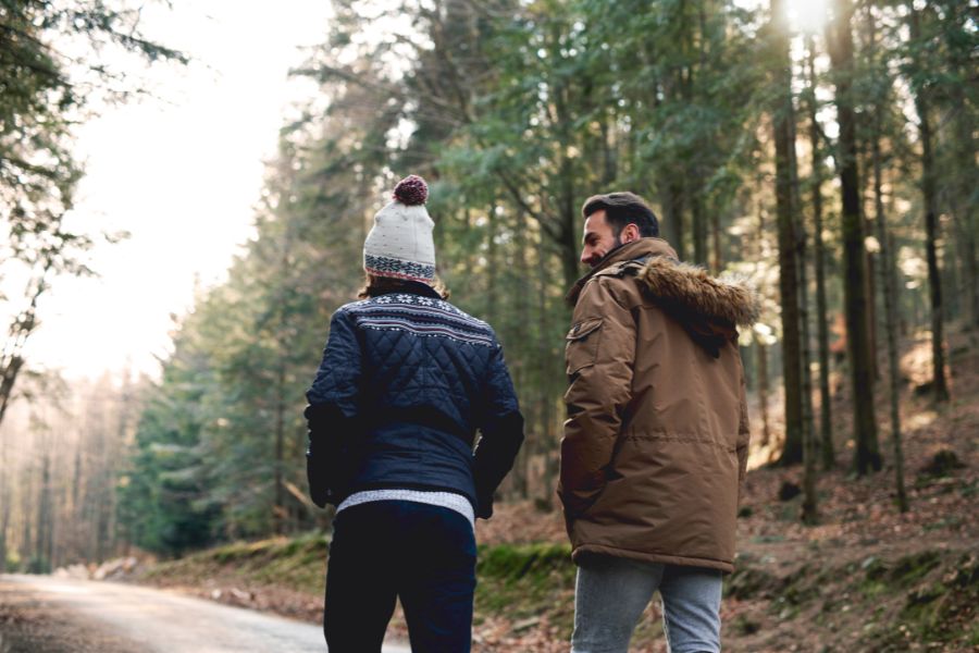 Två personer promenerar sida vid sida i en skog, klädda i varma vinterkläder, med en stig omgiven av träd i bakgrunden