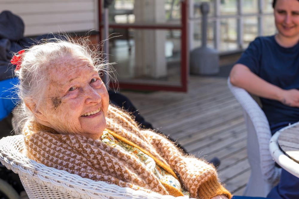 Äldre kvinna med stickad sjal ler under en solig dag på en uteplats, sittande i en vit rottingstol. En yngre person sitter i bakgrunden