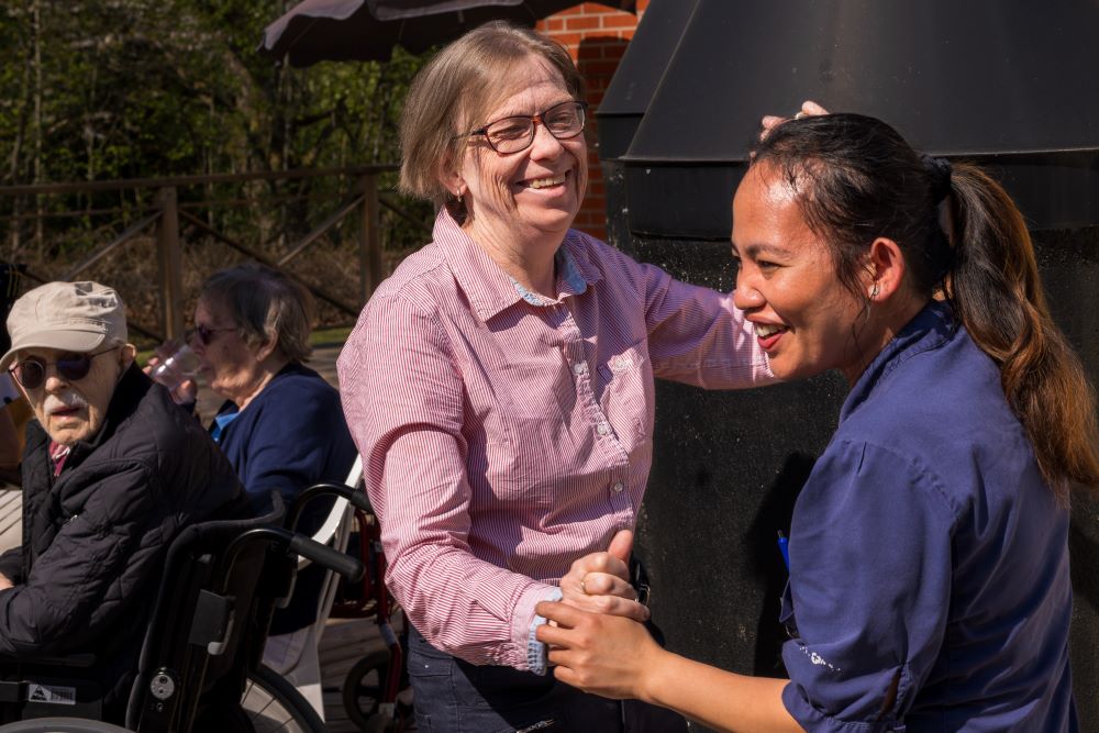 Personal och boende som ler och interagerar glädjefullt utomhus på en solig dag, med äldre människor i bakgrunden som sitter i rullstolar