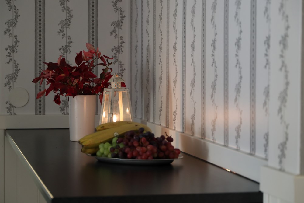 Blombukett i vit vas och fruktfat med bananer och vindruvor på mörkt träbord, bredvid en tänd lykta, mot en blommig tapet