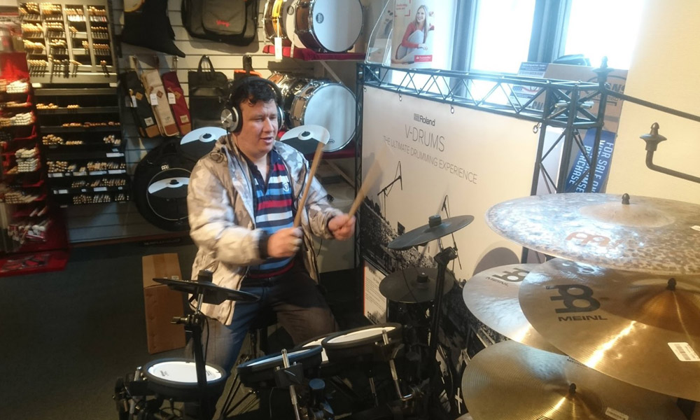 En boende spelar på ett elektroniskt trumset, omgiven av trumtillbehör och trumskinn. Han bär hörlurar och använder trumstockar