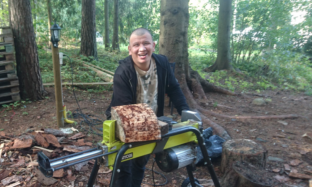 Man som använder en vedklyv i skogen, omgiven av träd och bark, under en klar dag