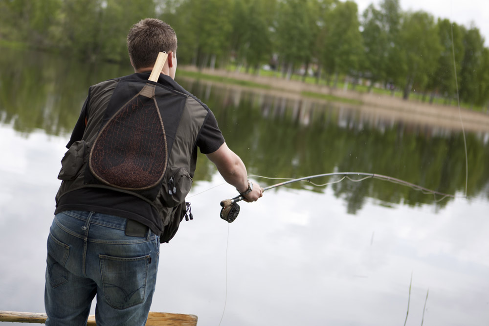 Person som fiskar vid en sjö med kastspö och håv på ryggen, omgiven av grönskande natur
