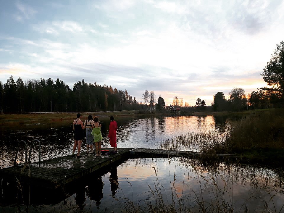 Fyra personer står på en brygga vid en sjö i solnedgången, omgivna av skog och lugn natur 