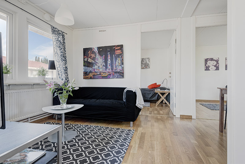 Ljust vardagsrum med svart soffa, färgglad tavla på väggen, träparkettgolv och stort fönster