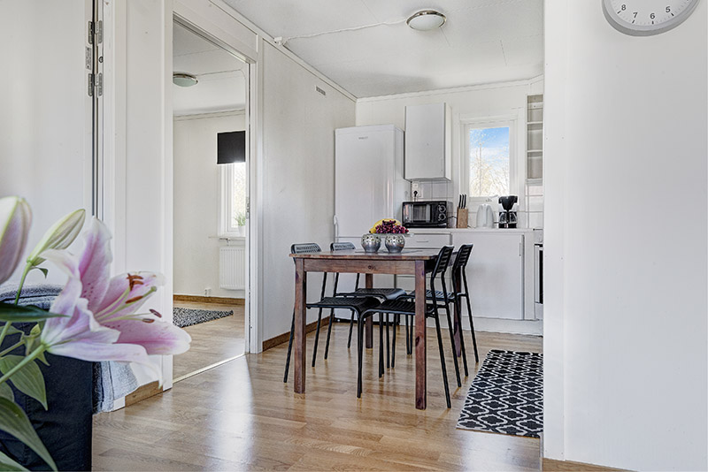 Ljust kök med matplats och fyra svarta stolar, vita köksskåp och trägolv, dekorerat med blommor och modern inredning