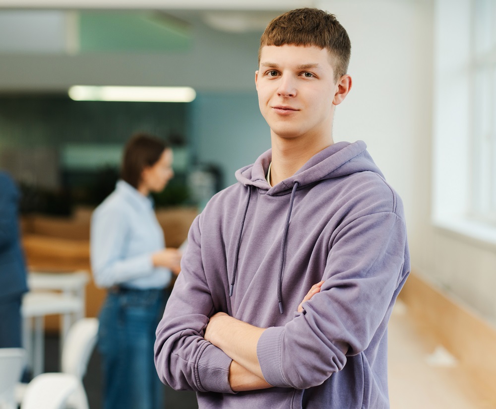 Ung man i lila hoodie med armarna i kors, står i skolmiljö med suddiga personer i bakgrunden