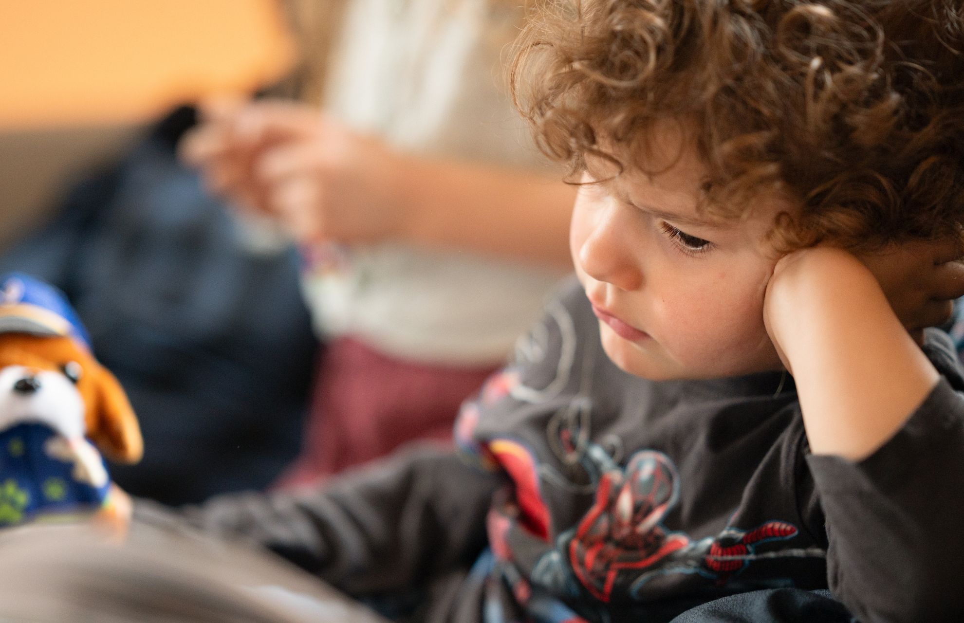 Litet barn med lockigt hår sitter tankfullt med huvudet lutat mot handen, en leksakshund syns i förgrunden