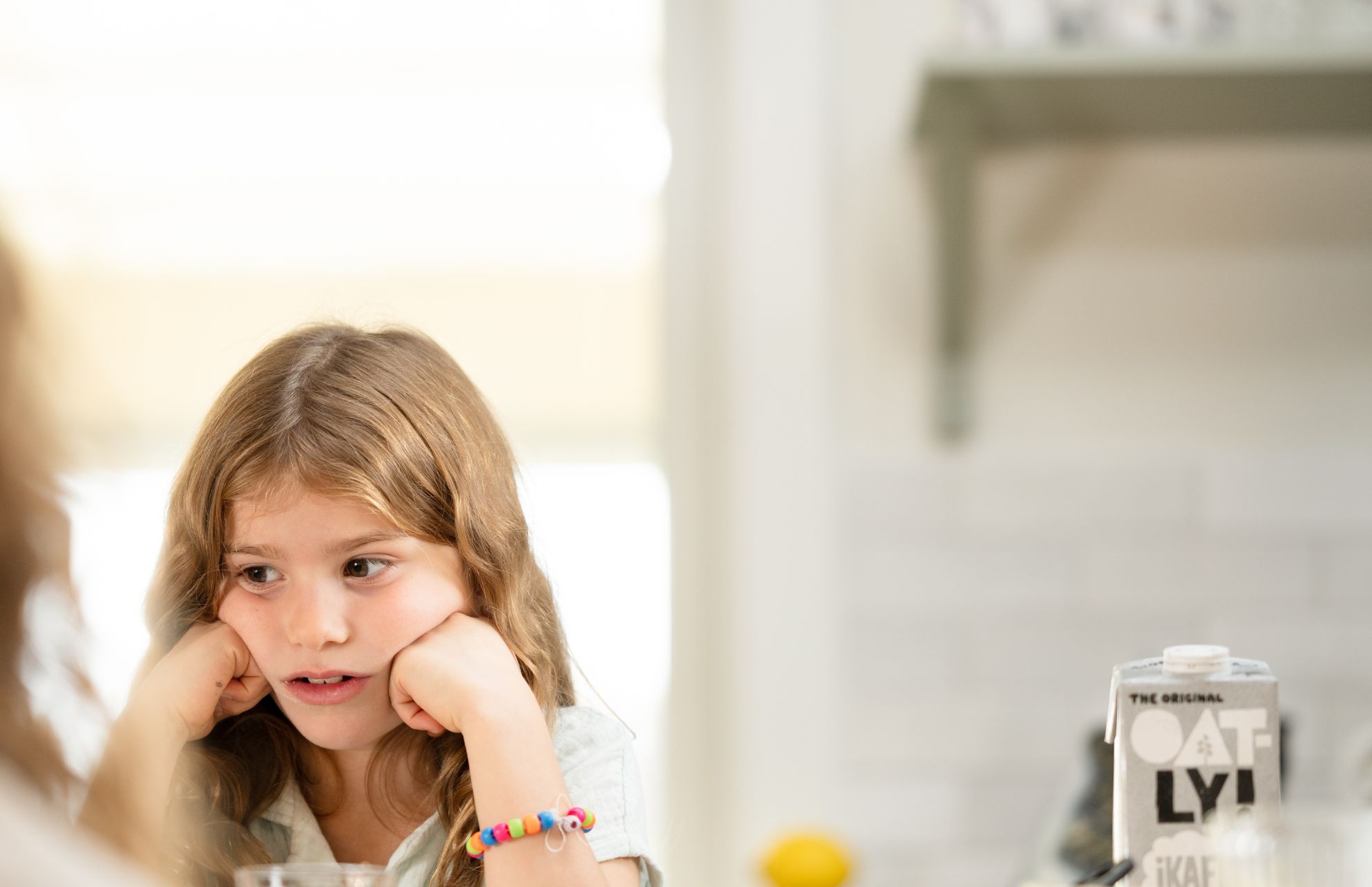 Tre personer sitter runt ett köksbord och äter frukost. Bordet är dukat med skålar, en kaffekanna och mjölkförpackning. Lampan är av glas och grön. Stämningen verkar glad och avslappnad