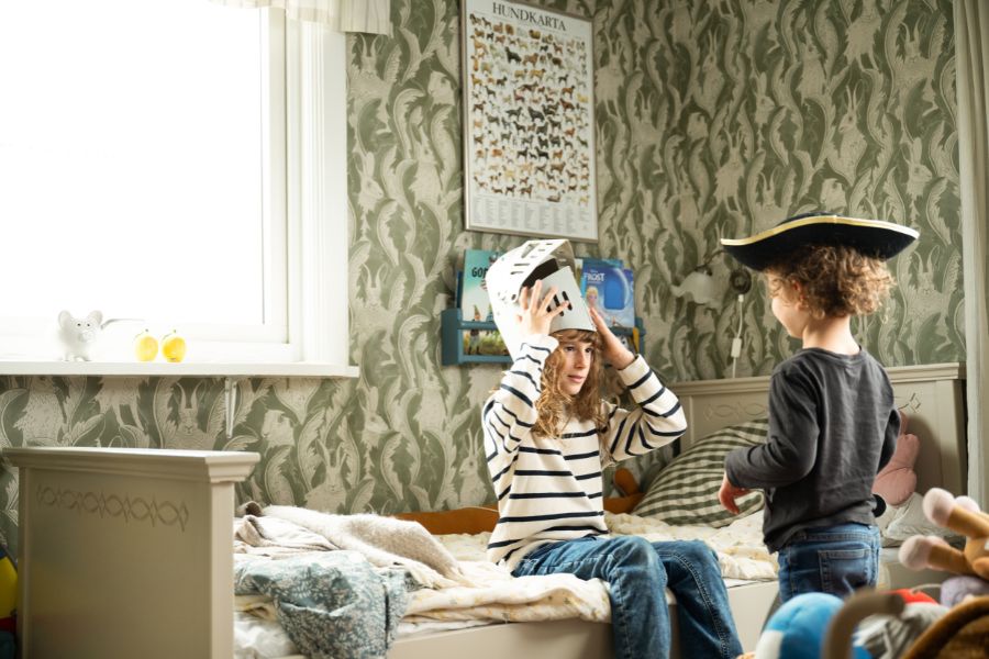Två barn leker pirater i ett sovrum med mönstrade tapeter, ett av barnen bär en pirathatt
