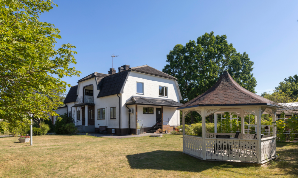 Vit villa med mörkt tak omgiven av trädgård, grönska och ett lusthus i trä på en solig sommardag