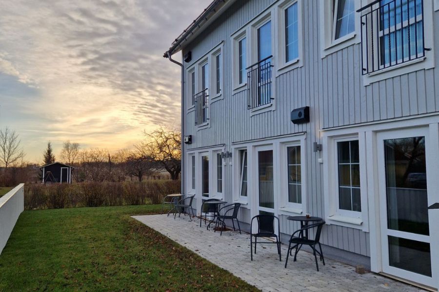 Tvåvånings hus i grått med en stensatt uteplats och stolar, omgiven av en grön gräsmatta och med träd och solnedgång i bakgrunden