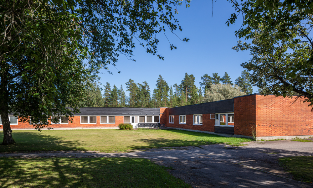 En låg byggnad i rött tegel omgiven av grönska och träd, med klar blå himmel i bakgrunden