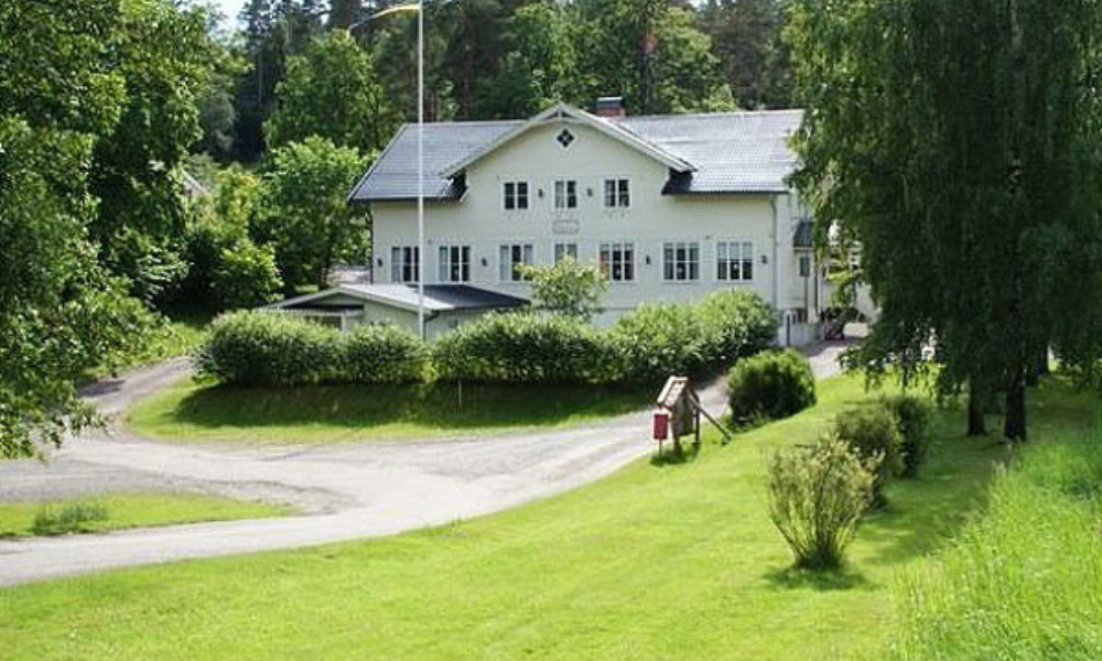 Vit byggnad i lantlig miljö omgiven av grönska och träd, med en grusväg som leder upp till entrén